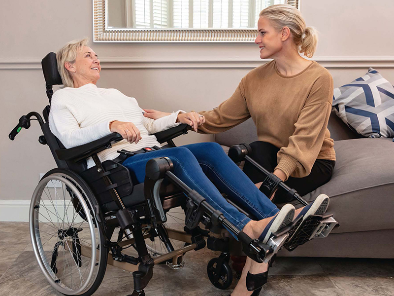 Frau sitzt in Multifunktionsrollstuhl neben einer Frau im Wohnzimmer